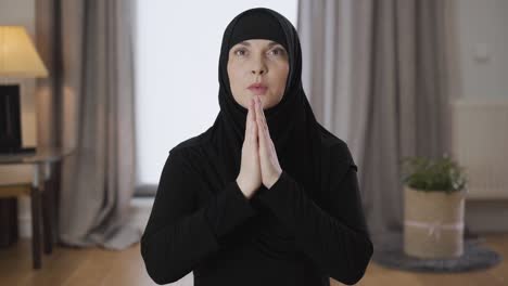 camera approaching slowly to face of young beautiful muslim woman holding hands together and praying. adult lady in black hijab talking with god at home. eastern culture, lifestyle, religion.