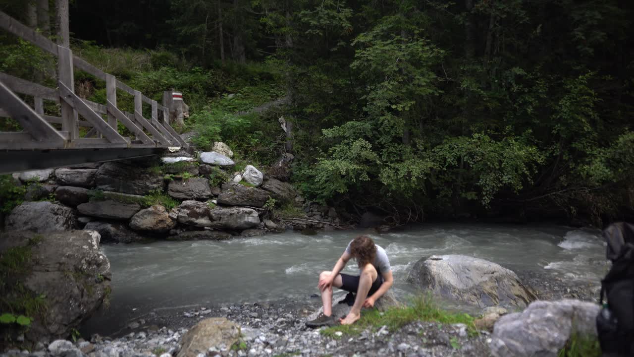 Escursionista stanco che riposa vicino a un fiume alpino nella foresta svizzera, alle prese con vesciche ai piedi dopo una lunga camminata, natura selvaggia remota, lotta realistica all'aperto, natura montana aspra.