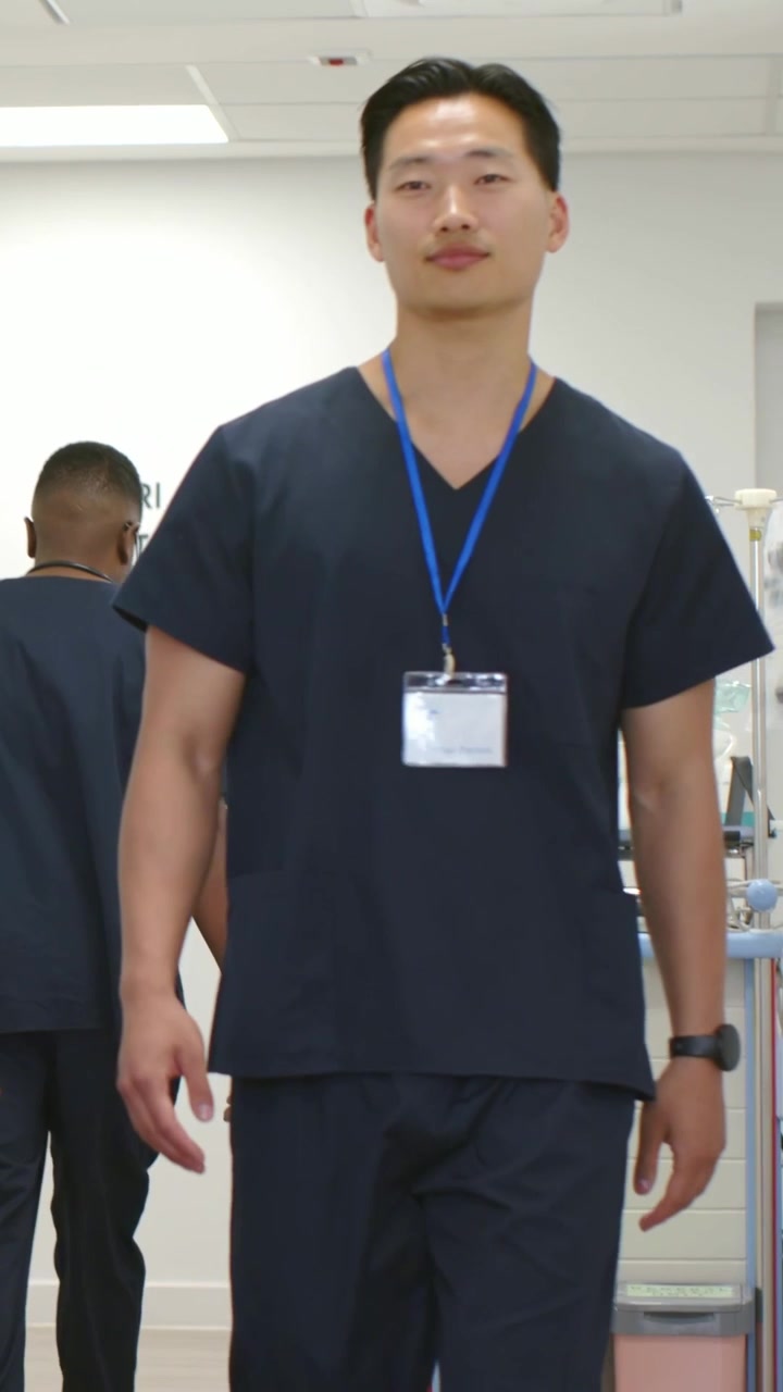Video verticale: Tecnico asiatico in camice che cammina dopo una consultazione, passando una sedia a rotelle verso il corridoio di diagnostica per immagini