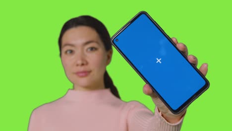 Studio-Portrait-Of-Smiling-Woman-Holding-Blue-Screen-Mobile-Phone-Towards-Camera-Against-Green-Screen-4