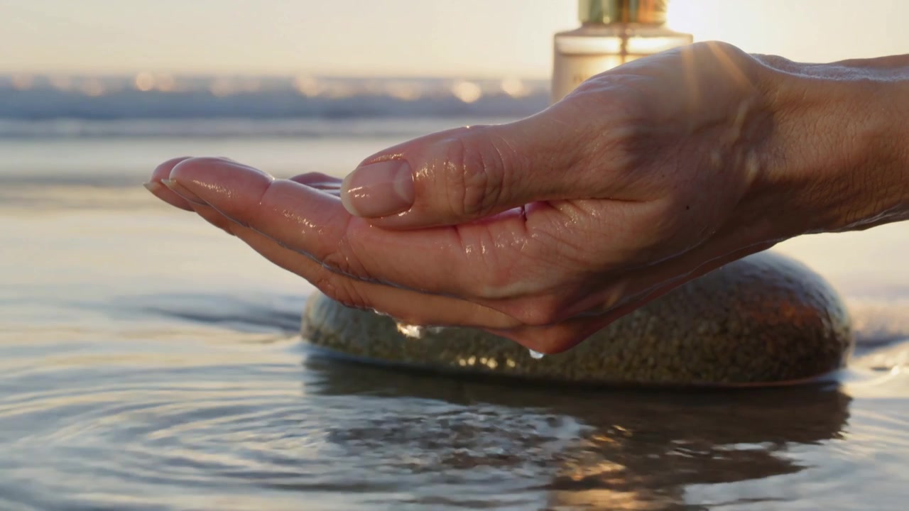 Primo piano di una mano che gocciola acqua su una spiaggia con una bottiglia di profumo al tramonto