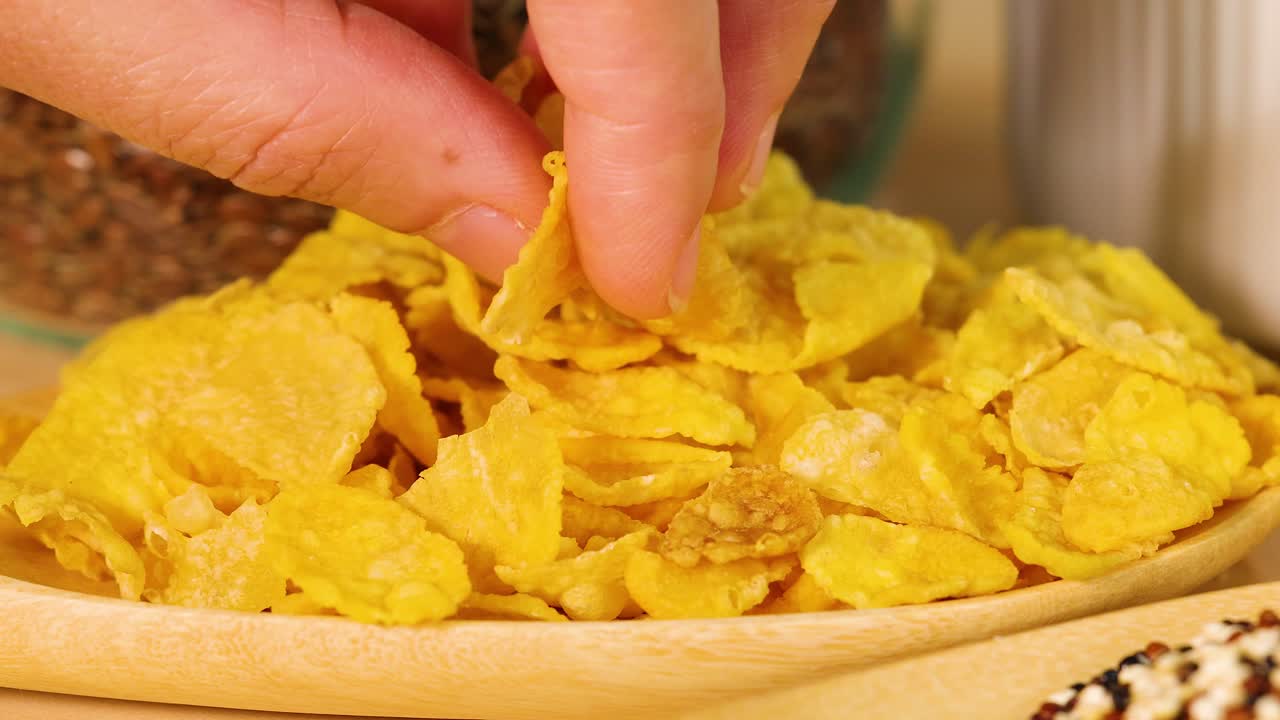 Una mano prende i cornflakes da una ciotola di legno in un ambiente cucina ben illuminato. I primi piani enfatizzano la consistenza e il colore