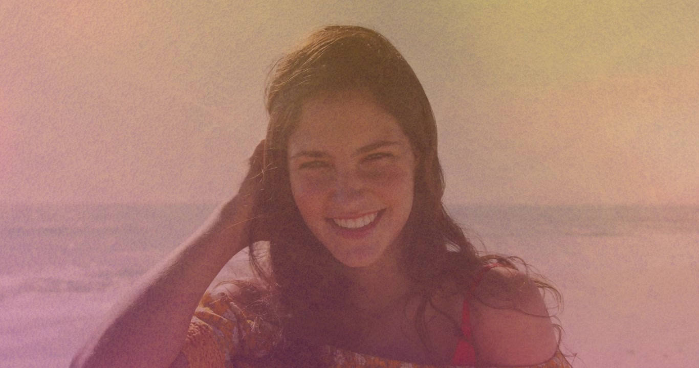 Donna in spiaggia che reagisce alla brezza, si sposta i capelli dal viso, sorridendo, con una sovrapposizione di grana cinematografica in stile moda.