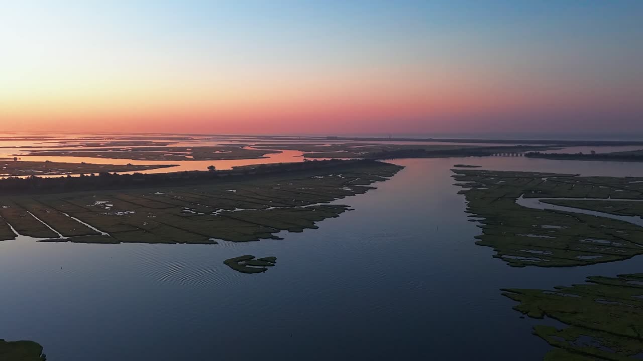 An aerial view high over the salt marsh off Freeport, NY during a colorful sunrise. The camera dolly in and slowly pan left over the waters and marsh grass, turning towards the sun