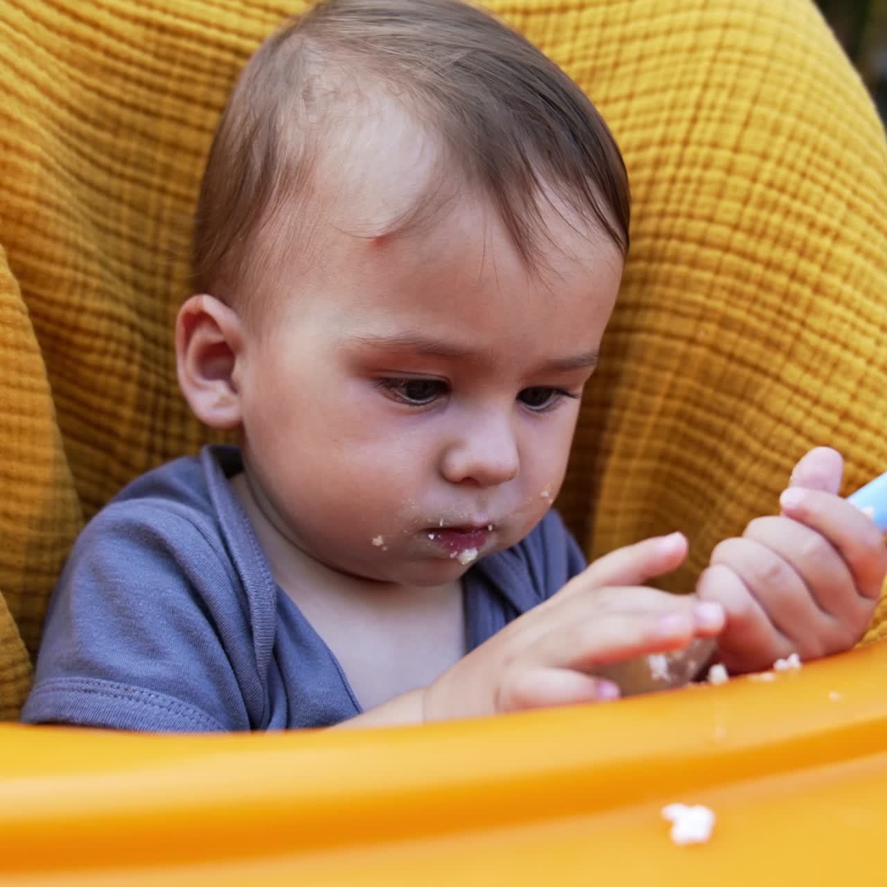 Caucasian baby boy sitting in yellow feeding chair. Little kid with smudged face after feeding turns spoon in hands and looking at it intently. Close up