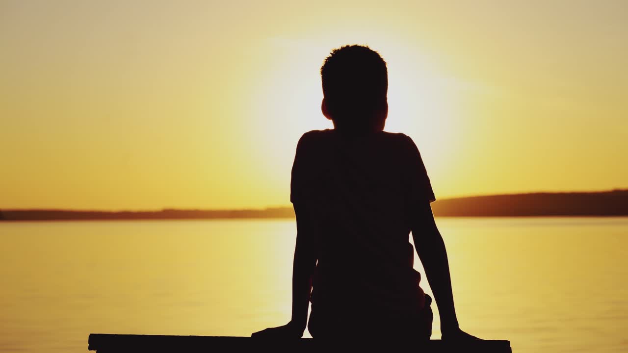Silhouette of a boy sitting on bench at beautiful sunset over the river. Child is watching the sunset and simulates flying near the lake