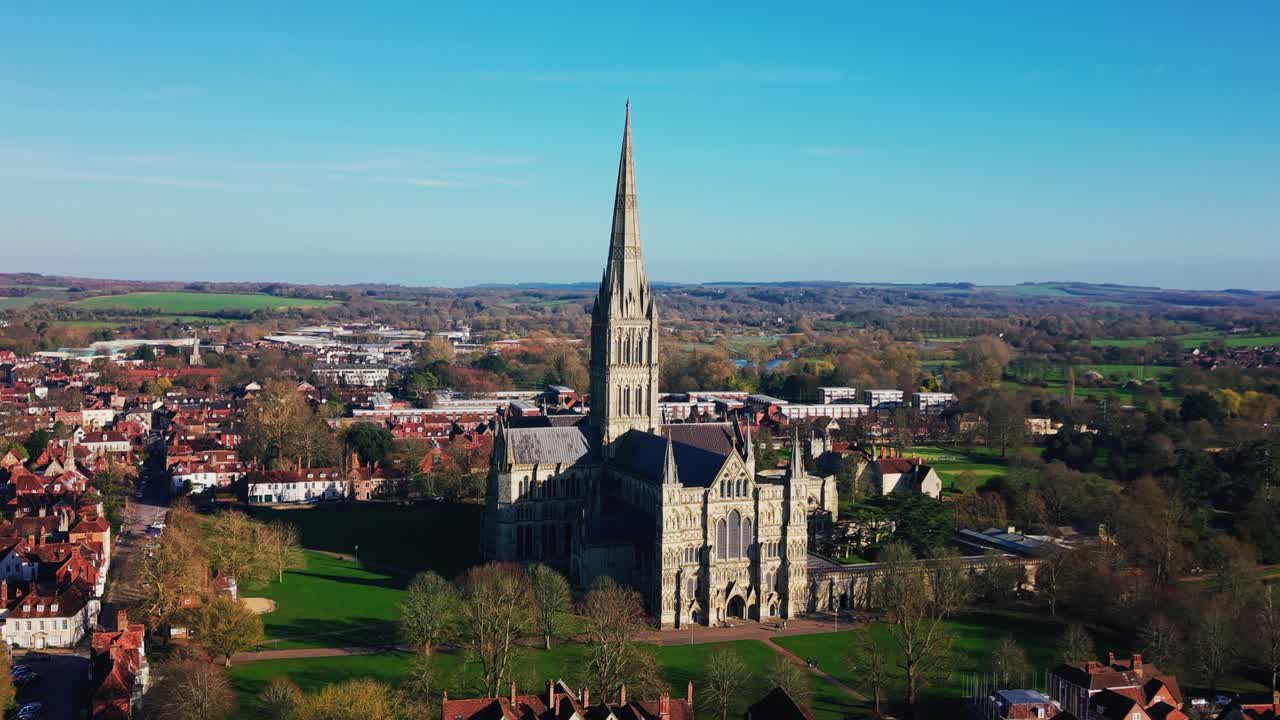 Kinowy widok z lotu ptaka na Katedrę w Salisbury i otaczającą panoramę miasta, ukazujący historyczną architekturę, zielone tereny katedralne i szerszą okolicę Wiltshire pod czystym, błękitnym niebem w Salisbury.