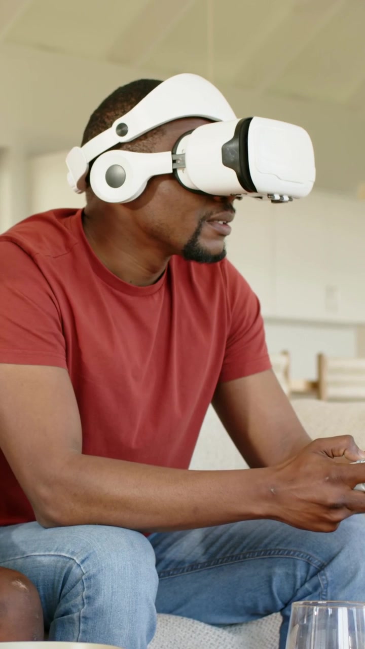 Vertikal video: Mand starter VR, læner sig på sofaen og spiller med controller derhjemme, iført headset og rød T-shirt