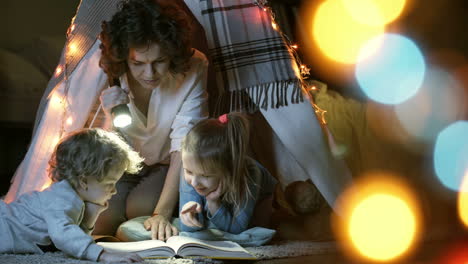 mother reads a book of fairy tales for her children while sitting in a tent at night