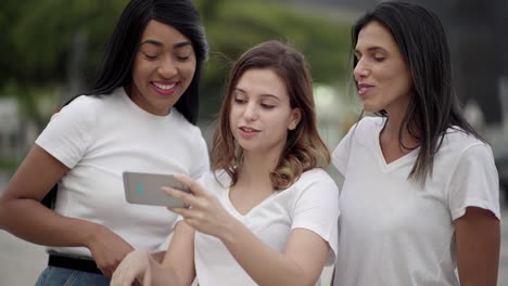 tres mujeres sonrientes de pie en la calle y mirando el teléfono