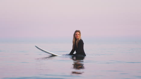 Mujer-Vestida-Con-Traje-De-Neopreno-Sentada-Y-Flotando-Sobre-Una-Tabla-De-Surf-En-El-Mar-Mientras-Las-Olas-Rompen-A-Su-Alrededor.