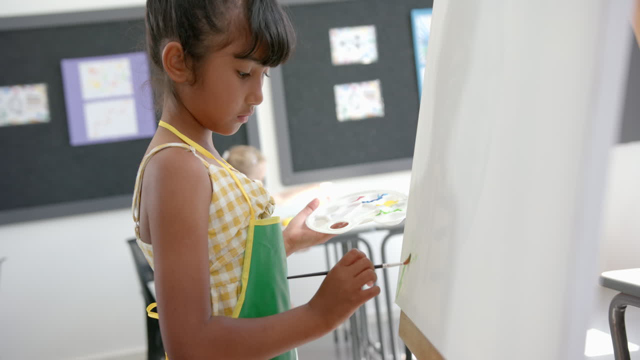 Biracial girl with a green apron is painting on a white canvas in a classroom at school