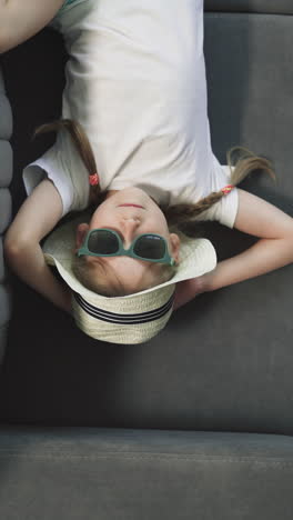 hand of mother taking suitcase while little daughter resting on sofa. cute preschooler girl in sunglasses and hat lies in hotel room upper close view