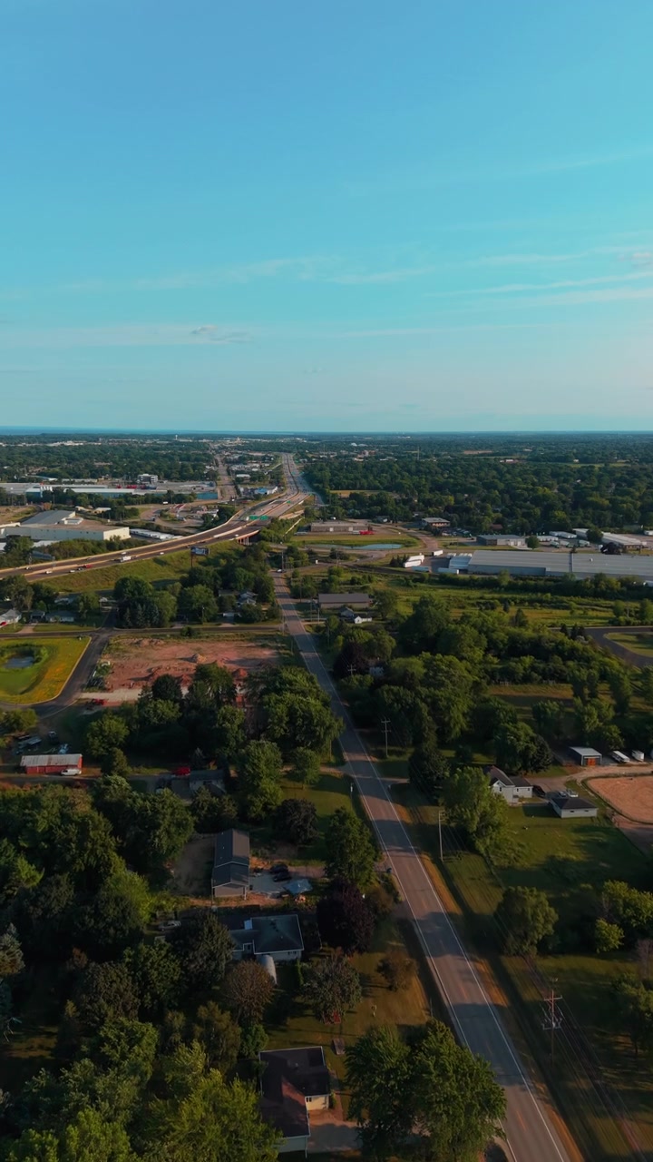 Suburban houses, trees, and a straight arterial road lead toward a highway under clear afternoon skies, useful as a calm small-town establishing shot or infrastructure b-roll.