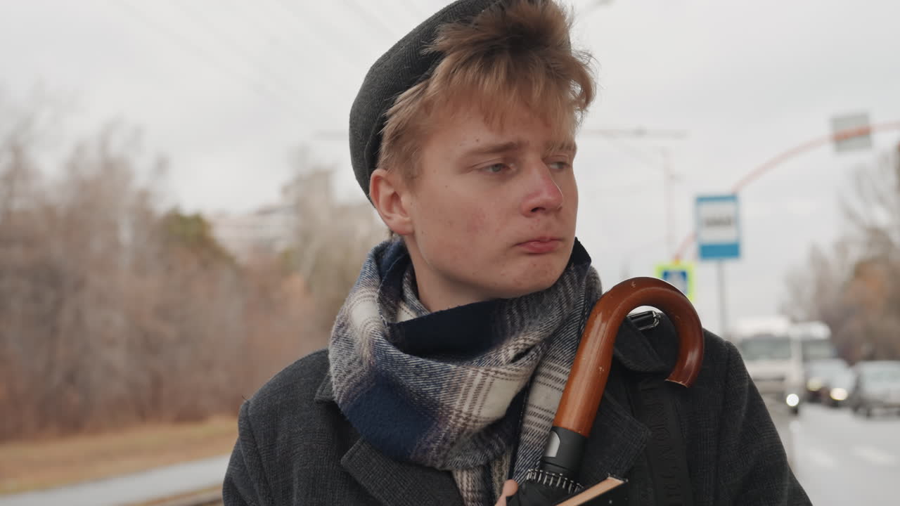 Young white creator writer at tram stop holding notebook and umbrella, bundled in heavy coat and patterned scarf, overcast sky, wet tracks, cars passing in distance, introspective gaze toward horizon
