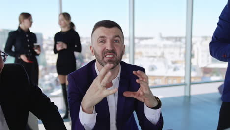 executive speaking emotionally on the camera and gesturing with his hands. two businessman sitting next to each other talking and smiling close up. two female office workers drinking coffee at the background.