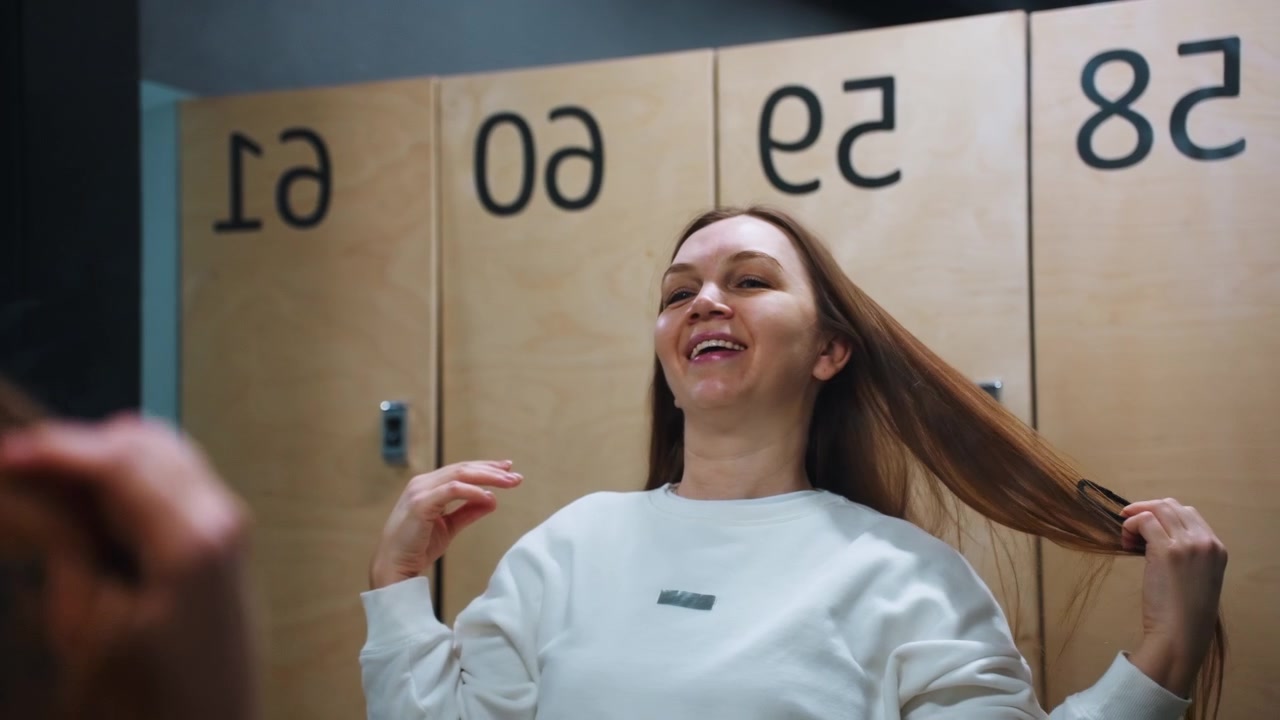 Donna caucasica sorridente nello spogliatoio si sistema i capelli mentre indossa una felpa bianca, armadietti di legno numerati sullo sfondo, espressione rilassata e postura sicura, routine di cura personale spontanea.