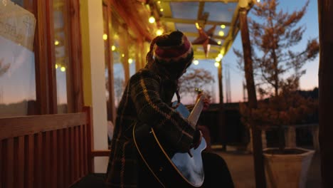 Guitarist-in-wool-hat-playing-at-dusk-under-patio-lights,-cozy-atmosphere,-side-view