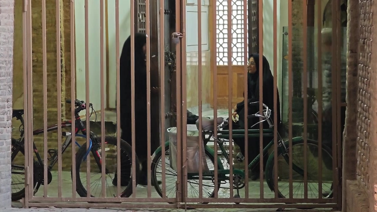 Donna con hijab seduta su una bicicletta dietro un cancello di metallo