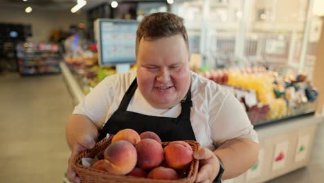 portrait: an overweight man at a supermarket worker in a white t-shirt and a black apron holds a basket of juicy peaches in his hands, smells them, smiles and looks at the camera