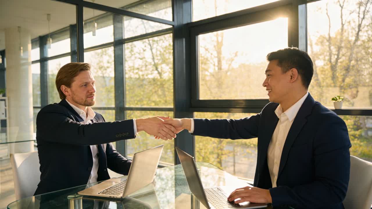 Confirming agreement with handshake, two men in suits moving to laptops in conference room with pen
