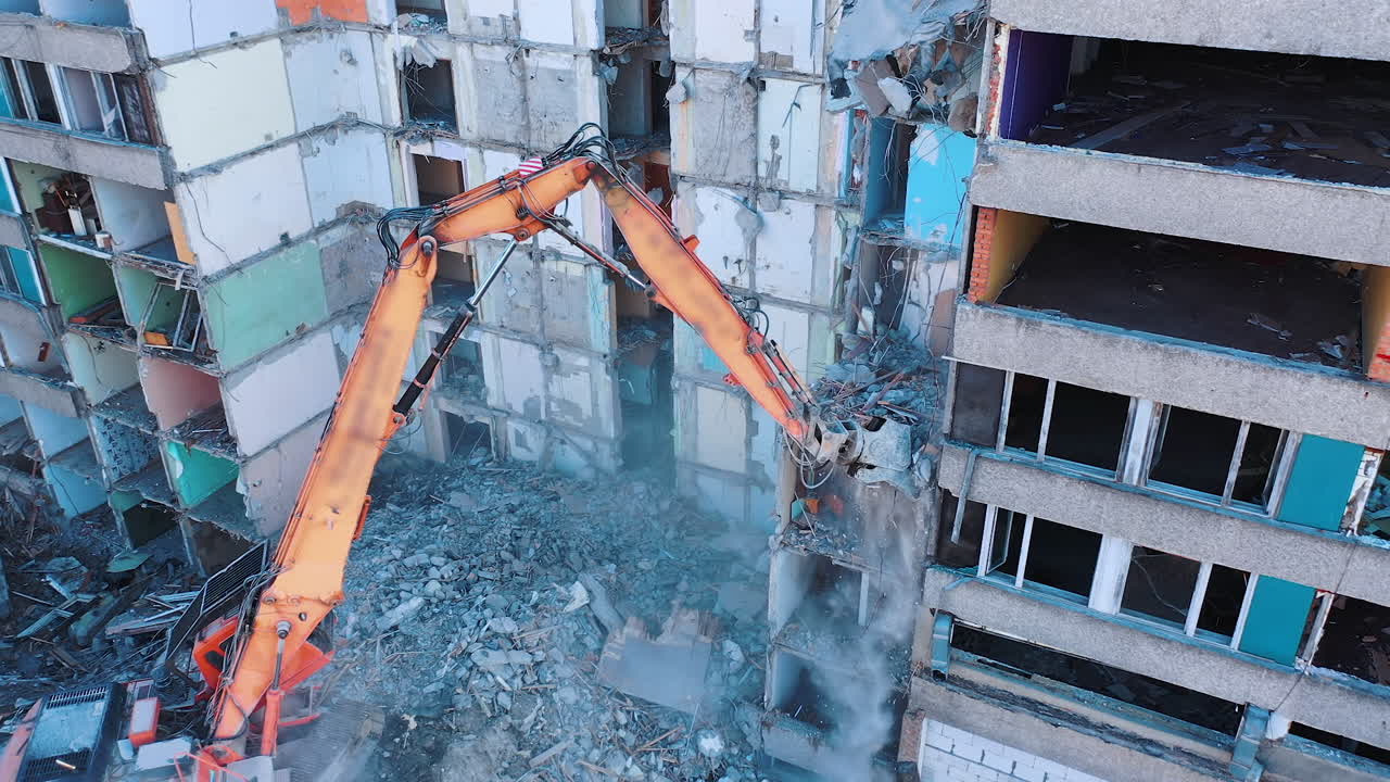 Ruined building is being demolished with the help of special machinery. Big claw is ruining the floors of the wrecked building. Debris falling and dust raising.