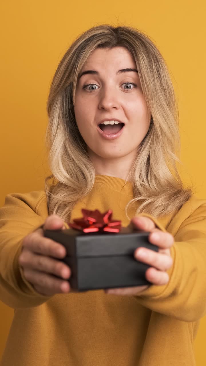 Happy young woman receiving gift box and saying thanks on yellow background