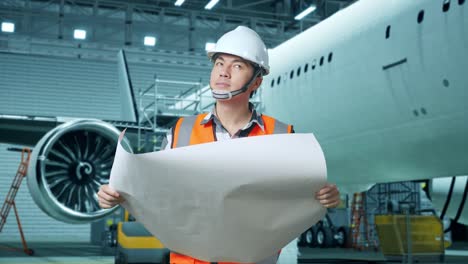 ingeniero masculino asiático con casco de seguridad mirando el plano en sus manos y mirando a su alrededor mientras está de pie con aviones en el hangar