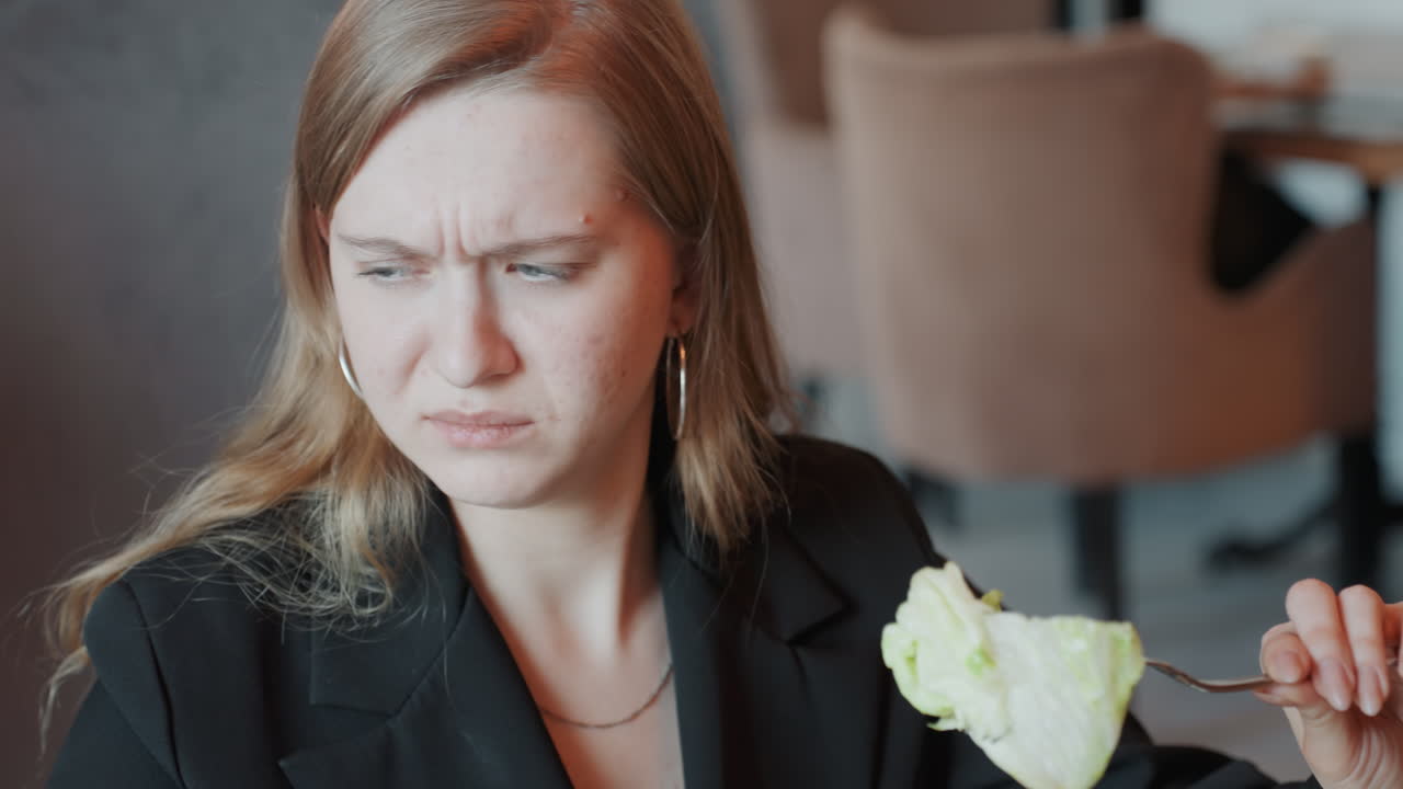 Donna che assaggia lattuga avariata in un caffè, manager d'ufficio in blazer ispeziona una foglia appassita sulla forchetta con una smorfia crescente, si copre la bocca con una reazione di disgusto, interno di ristorante discreto, primo piano spontaneo.