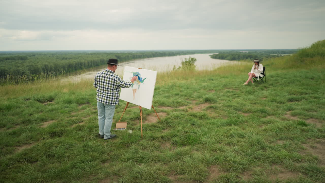 un artista, con un sombrero, una camisa a cuadros y pantalones vaqueros, se centra en crear una obra maestra en un lienzo en un campo de hierba exuberante al lado de un lago. una mujer con un sombreiro y un vestido blanco se sienta cómodamente en una silla