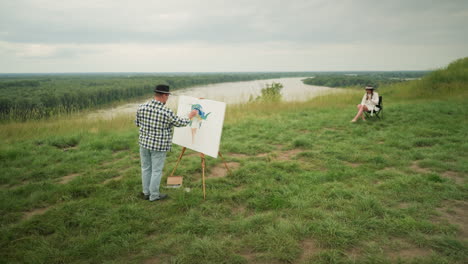 an artist, in a hat, checkered shirt, and jeans, is focused on creating a masterpiece on a canvas in a lush grassy field beside a lake. a woman in a hat and white dress sits comfortably on a chair