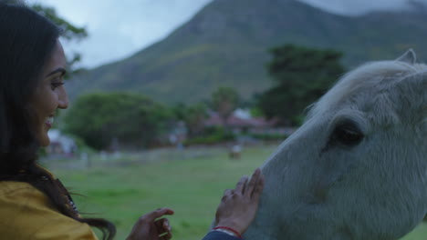 mujer joven india acariciando a un caballo sonriendo disfrutando de la vinculación cuidando a un animal doméstico amigo mostrando afecto en una hermosa granja rural