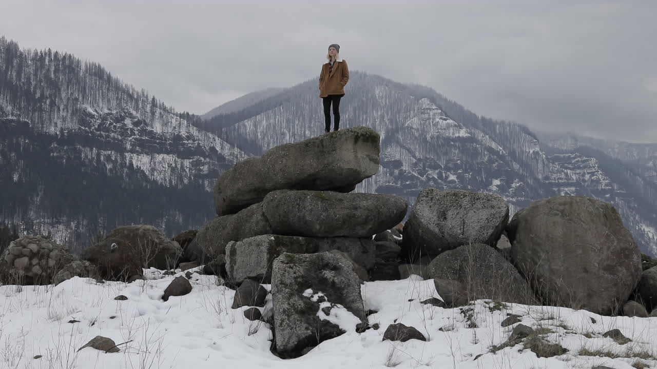 A girl stands on boulders with snowy mountains around her.