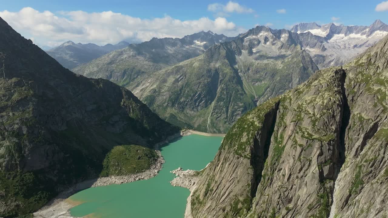 Alpine reservoir with turquoise water, lies between steep rocky cliffs, held back by a dam, towering mountain peaks rise in the distance, aerial shot