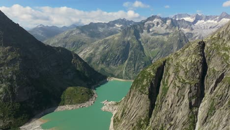 alpine reservoir with turquoise water, lies between steep rocky cliffs, held back by a dam, towering mountain peaks rise in the distance, aerial shot