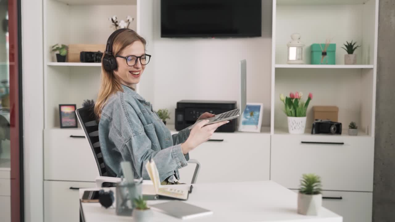 The young woman sits at her desk in her modern office and communicates via video conferencing through her laptop. She shows her new office through the webcam.She s wearing wireless headphones