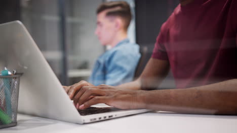 Close up of hands of young millennial man working at computer sitting in a glass office, close up, focus on foreground seen through glass
