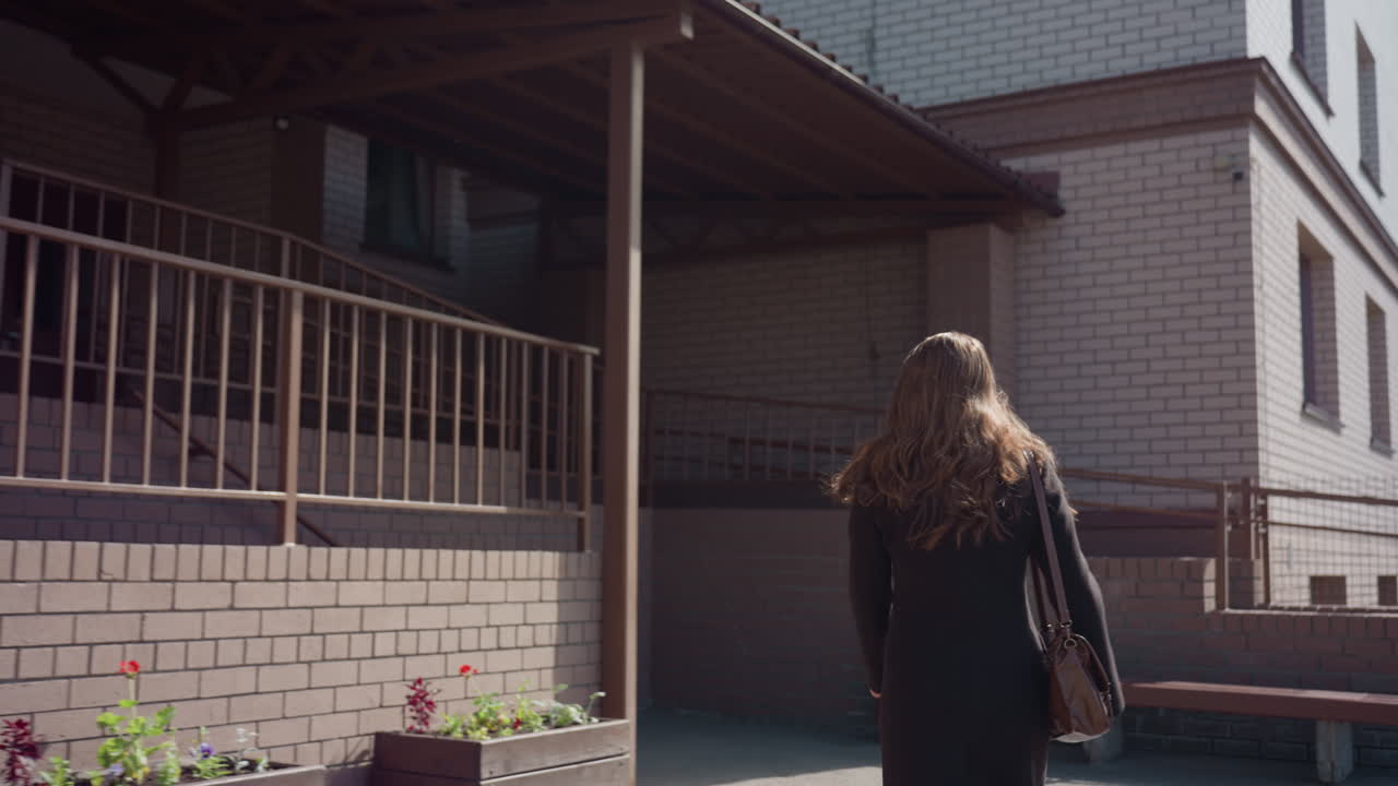 여성 인물이 벽돌 건물에서 안뜰로 나가고, 긴 머리가 흩날리며, 핸드백을 어깨에 메고, 덩굴로 덮인 격자 곁을 지나가요. 밝은 구름과 햇빛이 비치는 벽이 고요한 정오 분위기의 도시적인 외부 분위기를 만들어 냅니다.