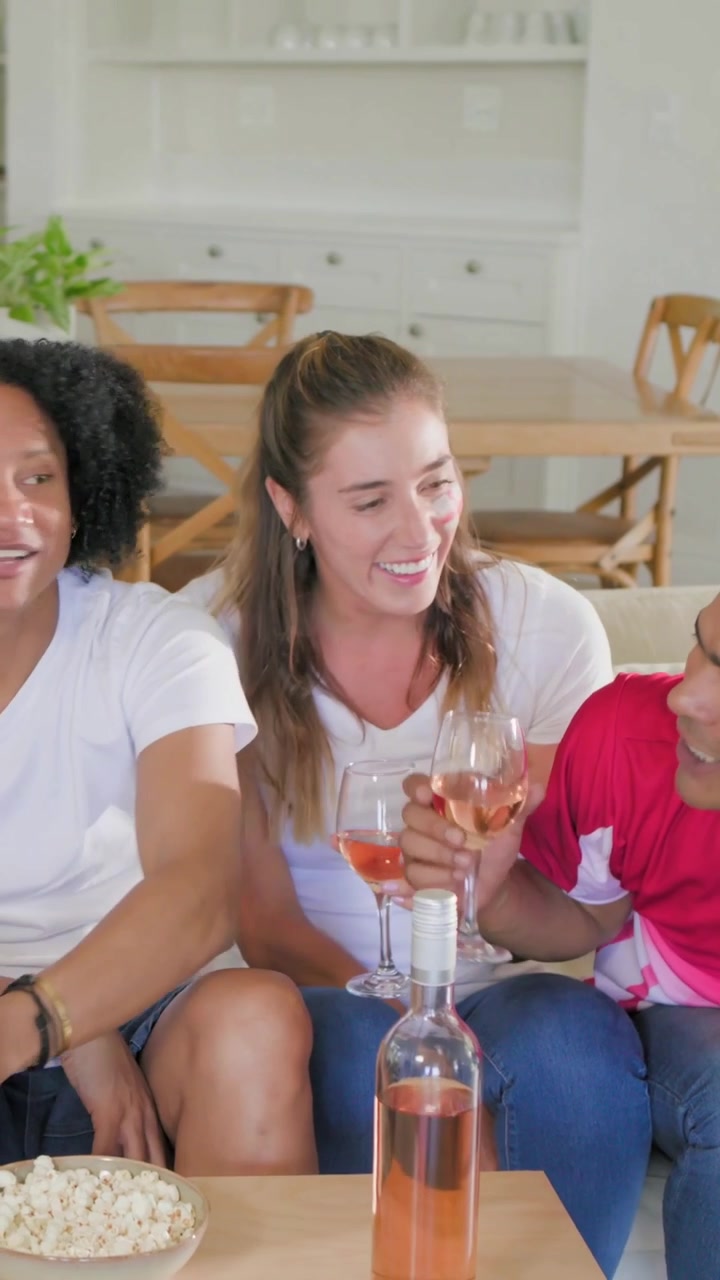 Vídeo vertical: Mulher inglesa e homem de camisa vermelha brindando com vinho rosé e uma bola, celebrando em casa.