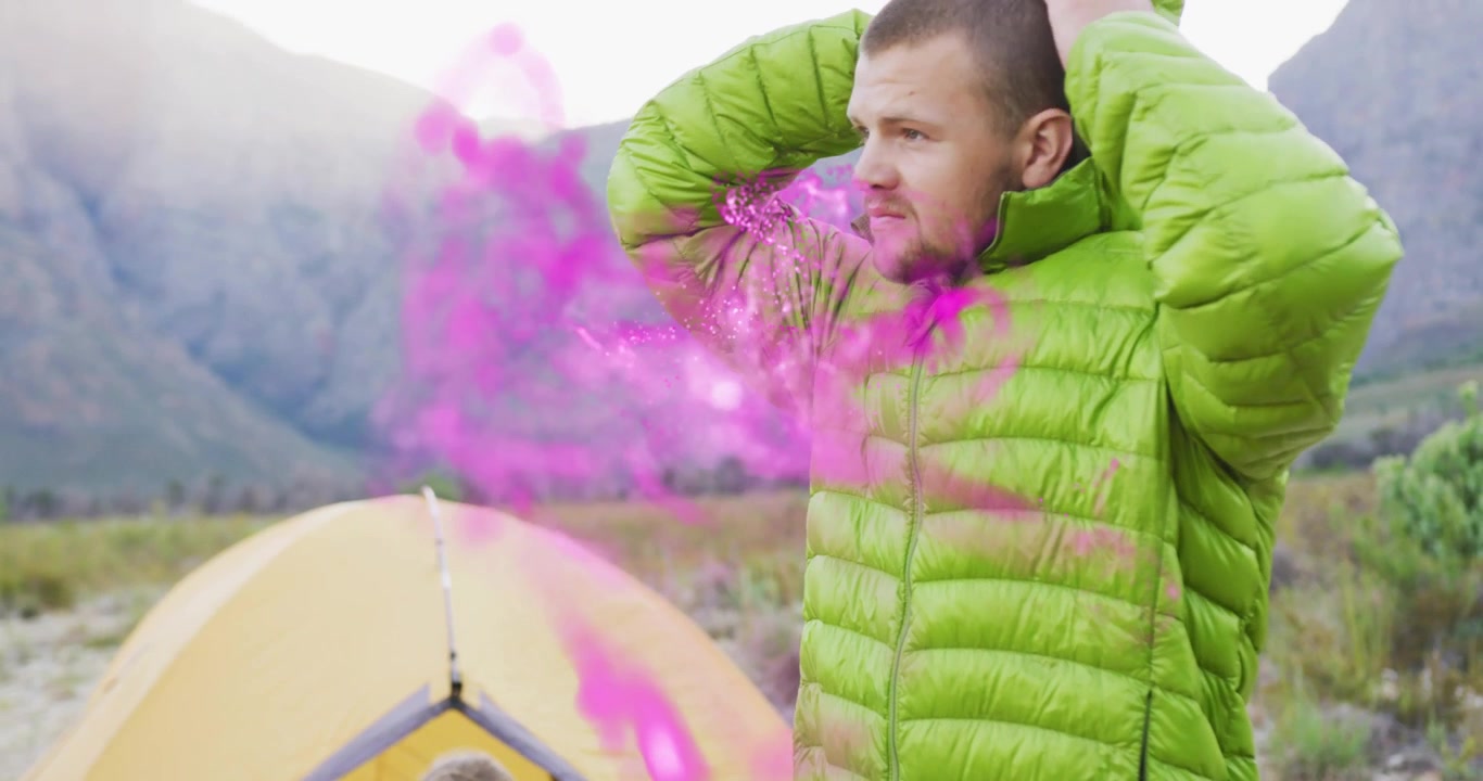 Uomo che si chiude la giacca verde e si protegge il viso da una nebbia rosa che passa vicino a una tenda gialla al campo.