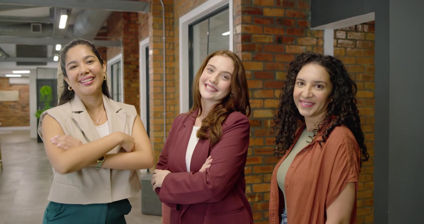 Equipo femenino diverso asintiendo y riendo mientras la mujer del centro comparte noticias en el pasillo de la oficina junto a una pared de ladrillo.