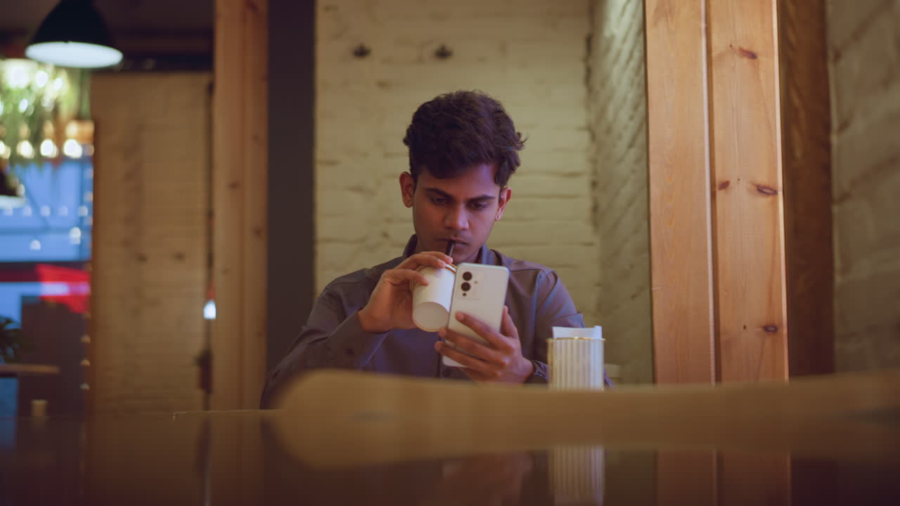 Young man in gray shirt sitting indoors at wooden table in cozy cafe, drinking from takeaway cup with straw while intensely focused on smartphone, surrounded by brick walls and warm lighting ambiance