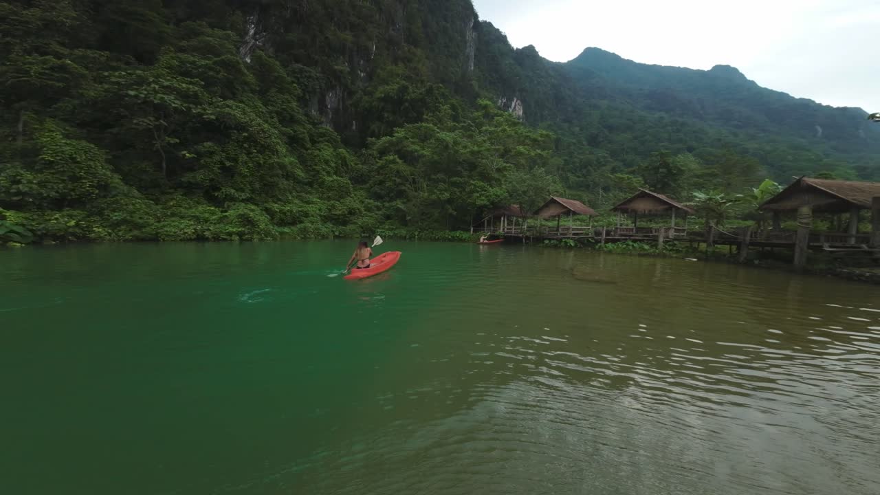 Scenic drone orbit of woman paddle boarding in water, Blue Lagoon’s vibrant green landscape in Vang Vieng, Laos, with blue waters and misty hills