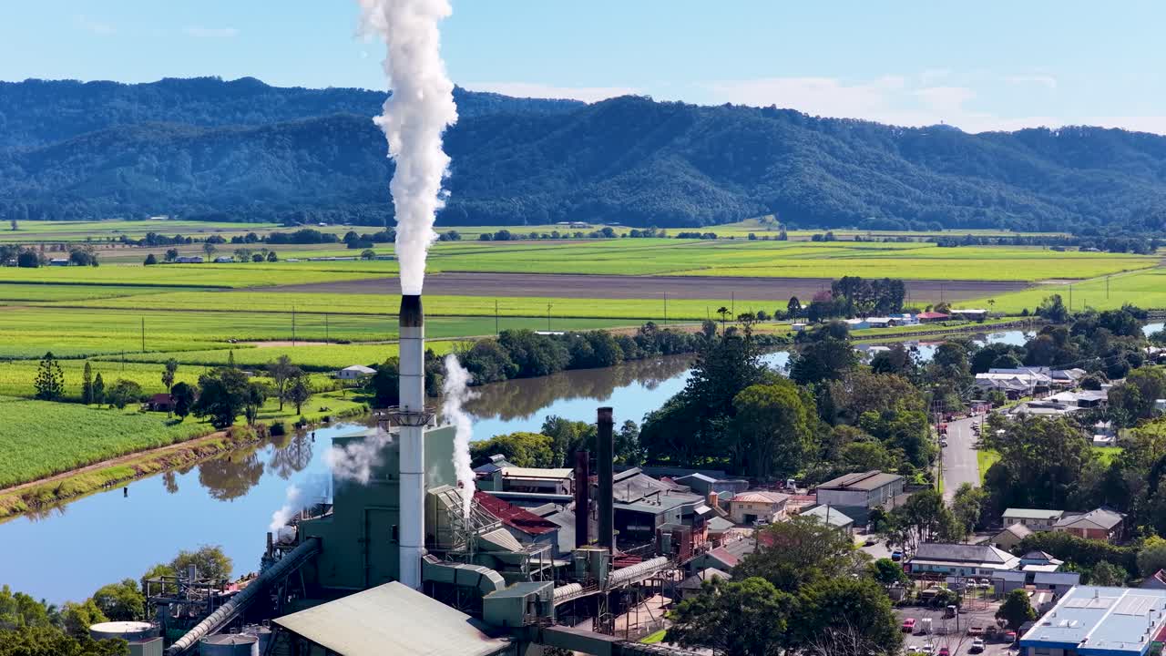 A factory emits smoke in a rural landscape with fields, river, and mountains under clear skies