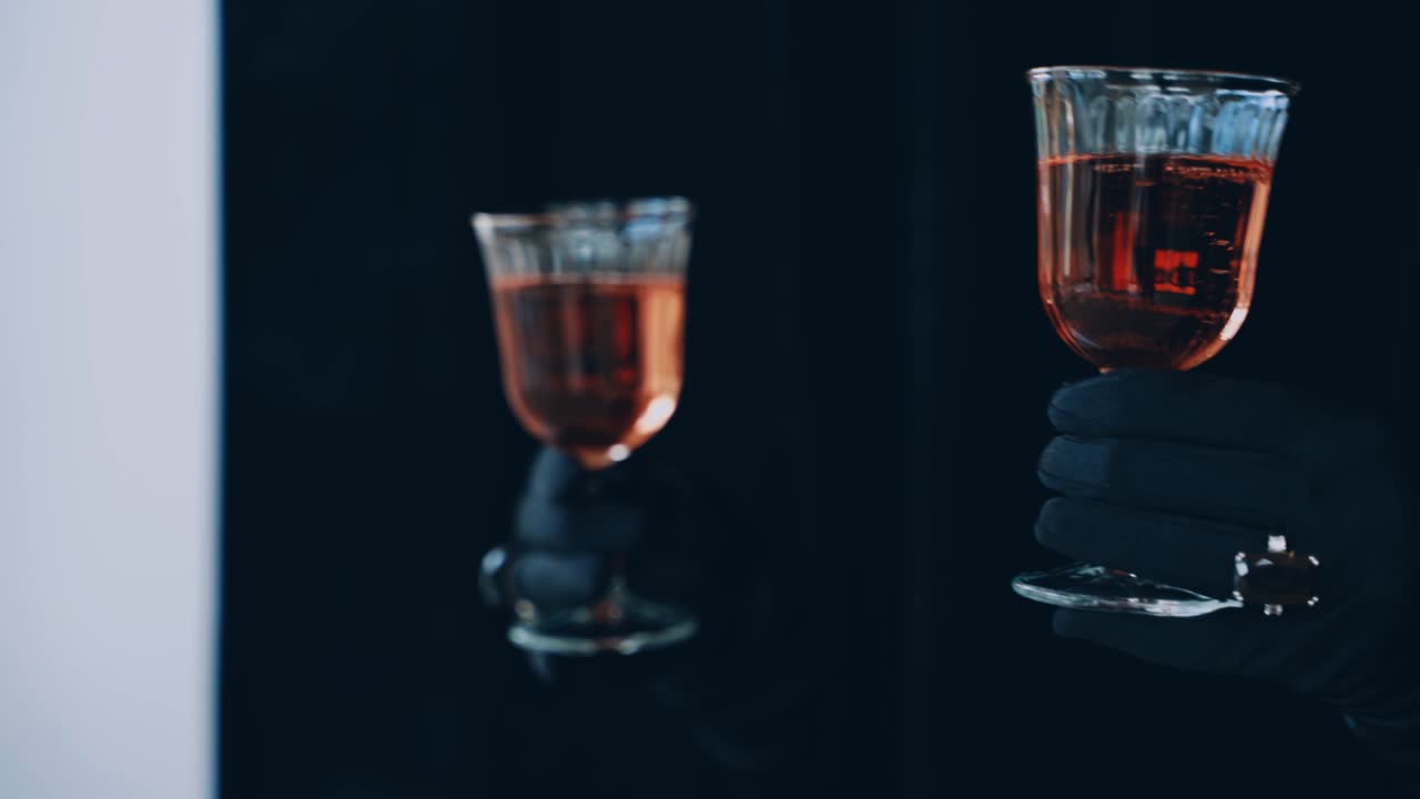 Two glasses of red champagne on the dark background. Enigmatic hands in luxury black gloves hold two glasses of wine.