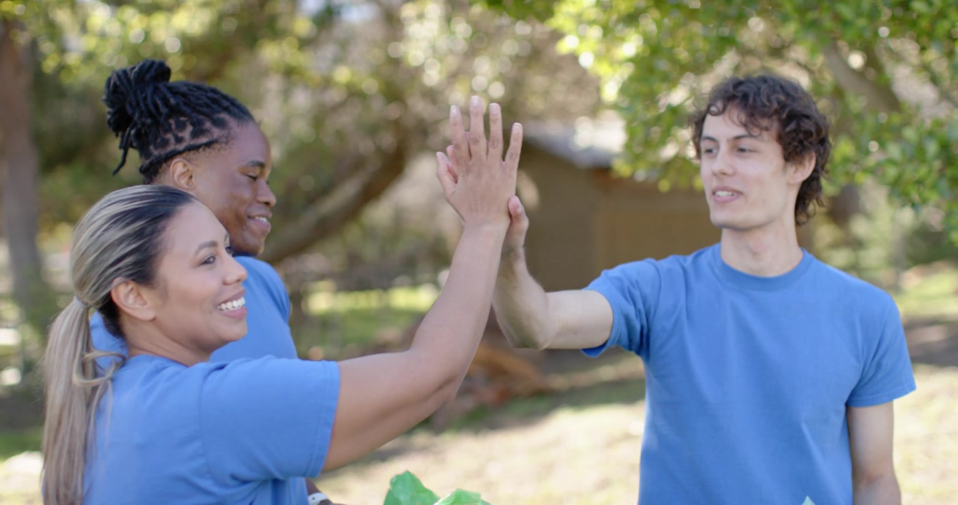 Diverse trio in blue tees with green bags right-hand man raising hand high-fiving cheering in yard