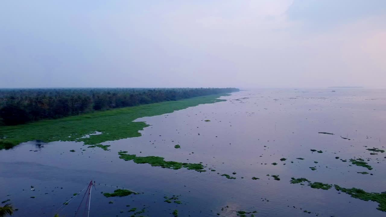 Luftpanorama dronebillede af Keralas floddelta ved solnedgang med traditionelle kinesiske fiskenet, frodige kokosplantager og roligt vand med flydende åkander i Indien