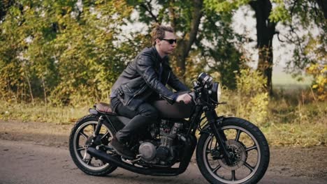 side view of a young man in sunglasses and leather jacket traveling with his bike on and asphalt road on a sunny day in forest in