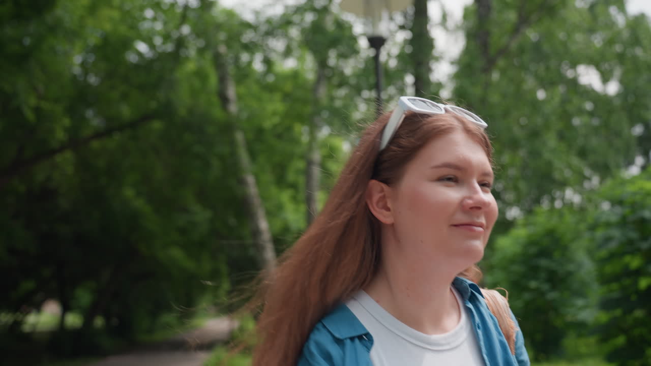 Young woman with long hair and white sunglasses smiles gently while walking through serene forest path, dressed casually in white shirt and blue jacket