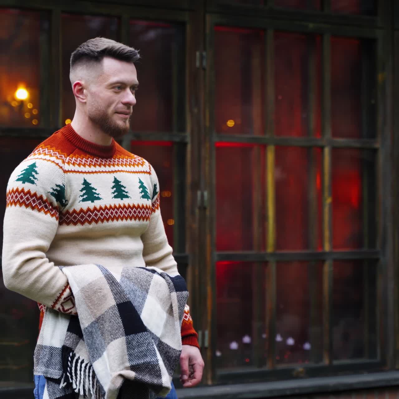 Smiling young guy on cottage background. Handsome man in warm sweater walking to someone in terrace. Guy with a warm blanket in hands outdoors.